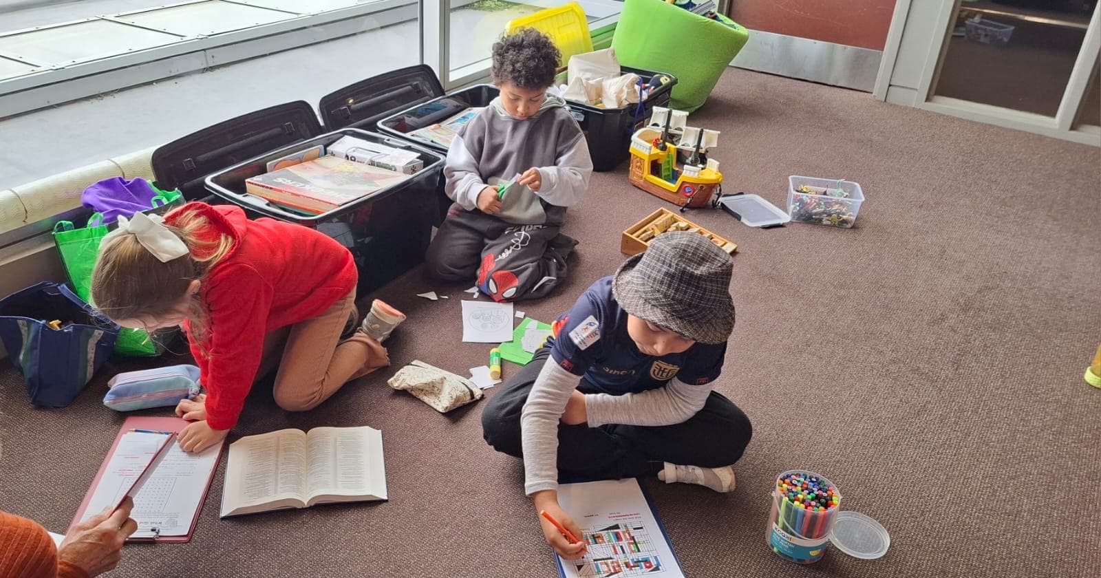 Children sitting on the floor by a window with Bibles, pens and craft boxes around them.