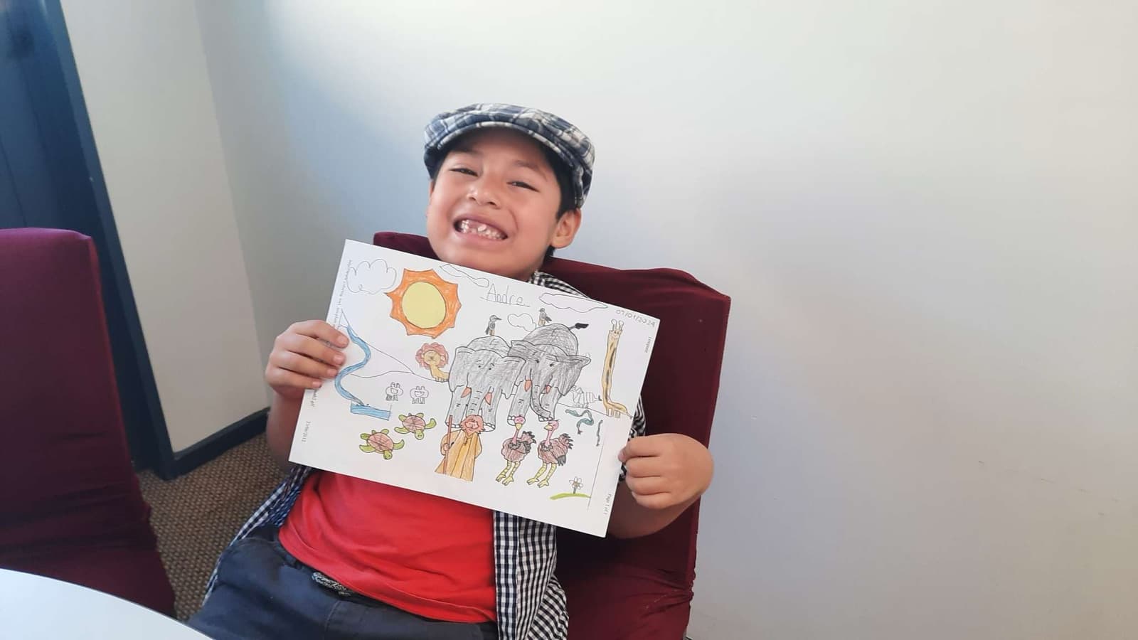 A boy in a flat cap grinning and holding up a colouring page he has finished.