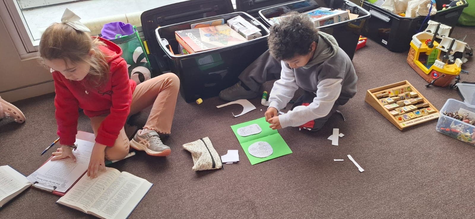 Two children on the floor, one writing in a workbook and one cutting out a paper shape.