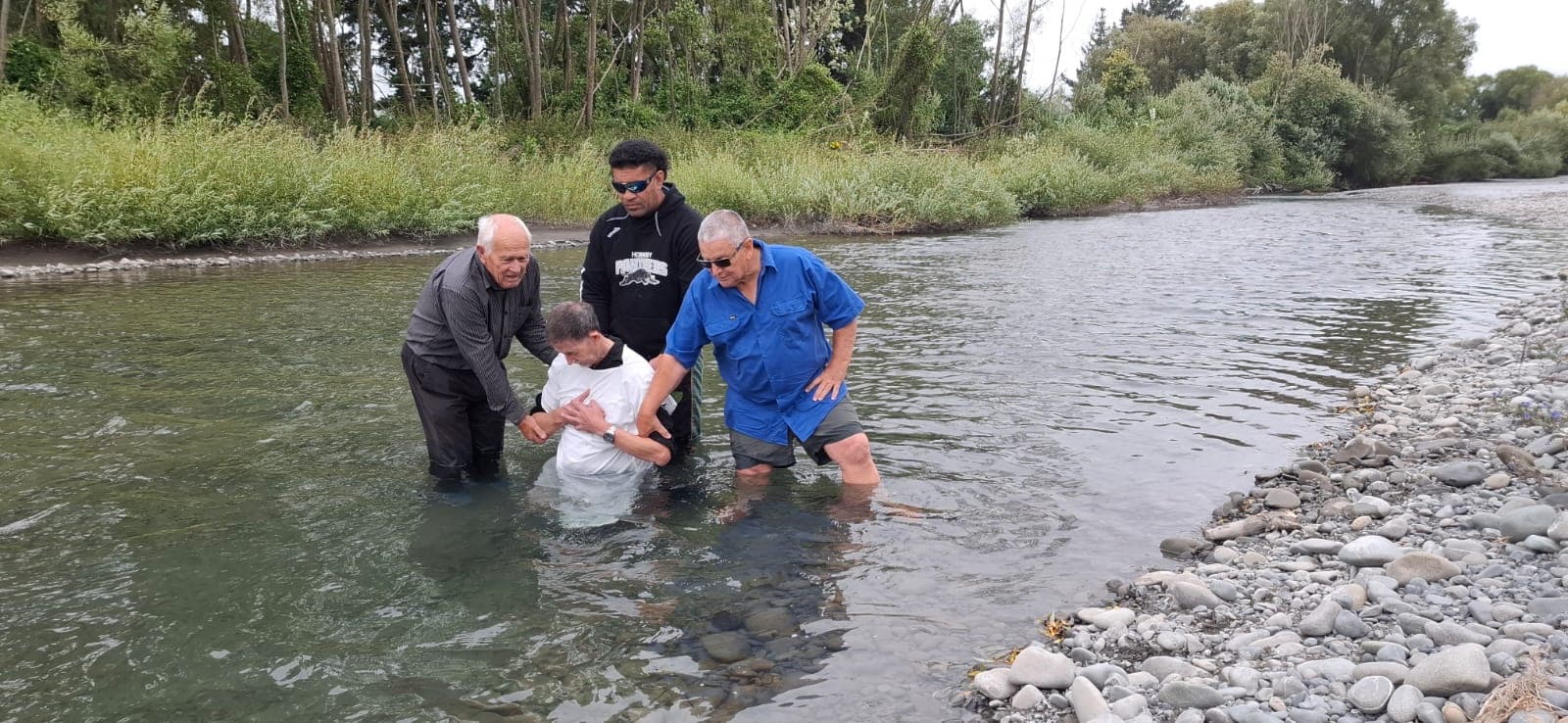 Three men standing in a shallow river, lowering a fourth person backwards into the water.