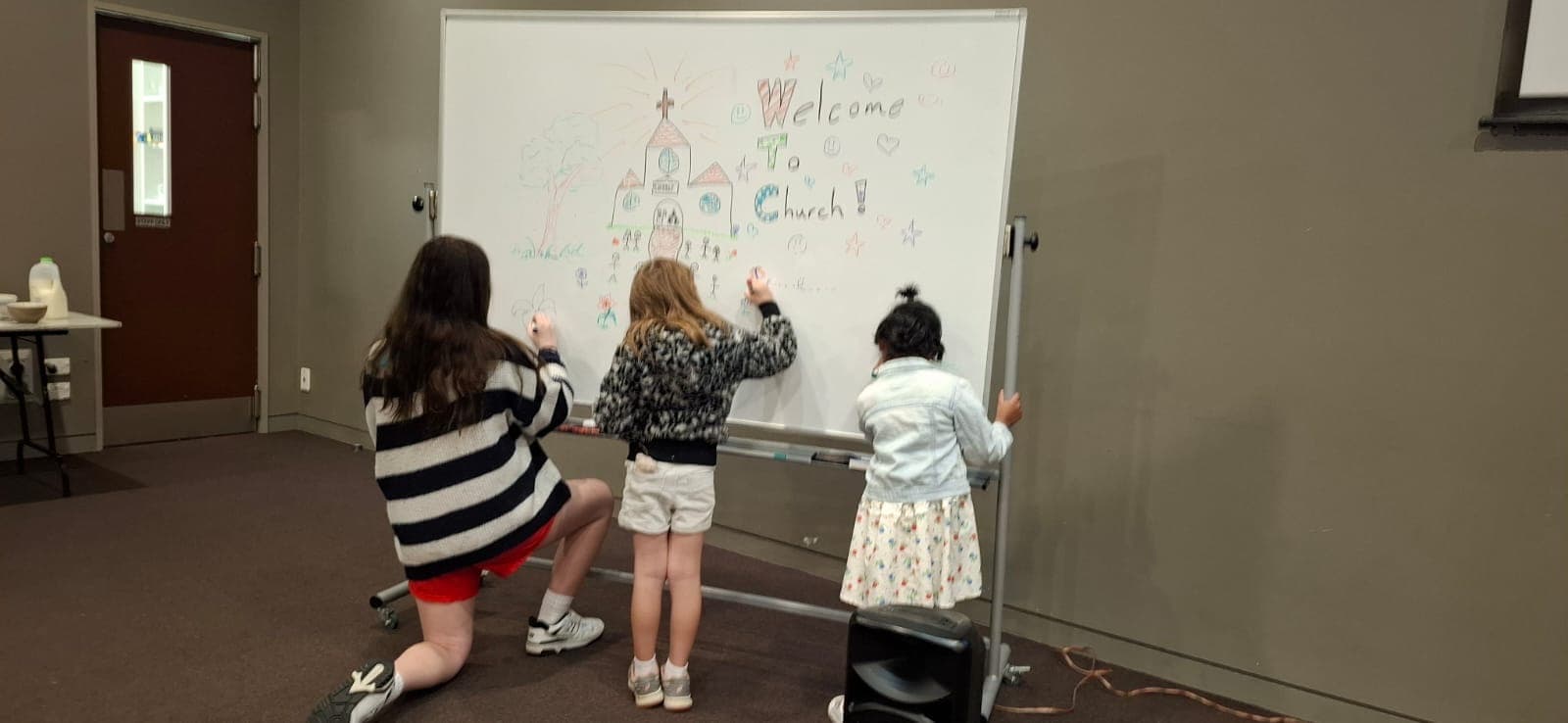 Three children, seen from behind, drawing a church and the words Welcome To Church on a whiteboard.